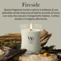 Una lampada da caminetto - Candela Media Woodwick, di colore bianco, con una fiamma tremolante, è adagiata tra legni trasportati dal mare, pietre e muschio. Il testo in italiano sopra descrive le note olfattive: bergamotto italiano, ambra dorata e mogano affumicato.