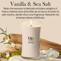 Una candela Woodwick alla vaniglia e sale marino - Candela Grande poggia su una superficie sabbiosa circondata da orchidee, foglie verdi rigogliose e pezzi di legno galleggiante. Il testo in italiano sottolinea la fragranza rilassante e sofisticata della candela: una miscela armoniosa di vaniglia cremosa e sale marino rinfrescante che evoca un'accogliente atmosfera costiera.