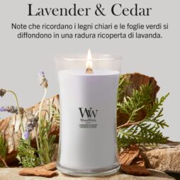 Una candela “Lavanda e Cedro” della linea Woodwick - Candela Grande, con cera color lavanda in un barattolo di vetro, poggia su una superficie di legno circondata da rametti di lavanda e foglie verdi. Il testo in italiano sopra descrive la sua fragranza come “Lavanda e Cedro”.