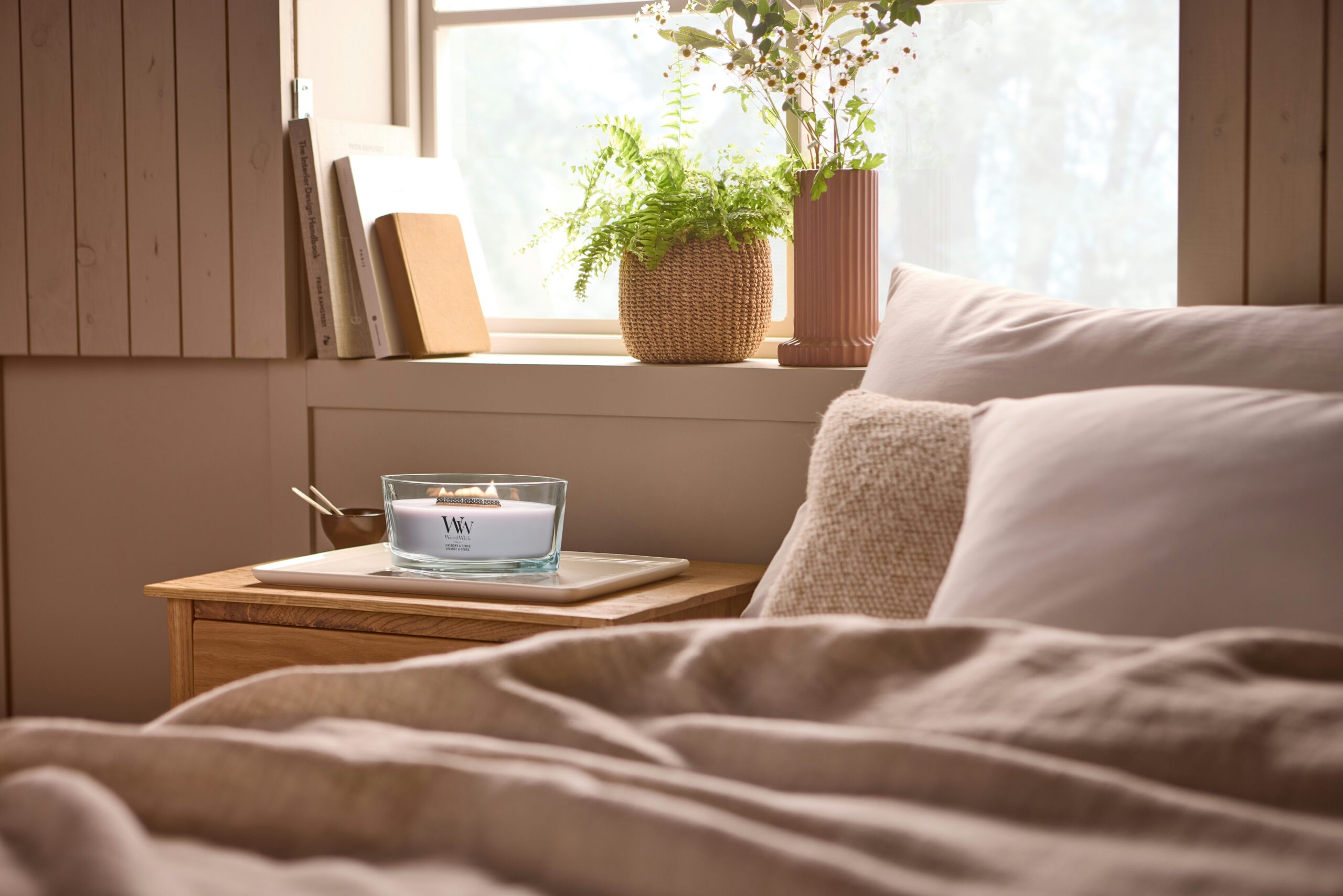 Lavender and Cedar – Ellipse – WoodWick Ellipse 5 Una camera da letto accogliente con un letto ben rifatto, una pianta, dei libri e la candela “Candela Ellipse Lavender and Cedar Woodwick” accesa su un comodino di legno vicino a una finestra inondata di luce solare.