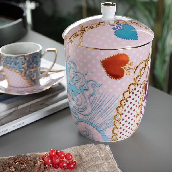 Contenitore in Porcellana con Coperchio – Henriette “Trend” Barattoli da Cucina - Salva Freschezza 2 La linea Scatola Trend - Henriette: un vaso decorativo in porcellana con coperchio, impreziosito da cuori colorati, dettagli dorati e motivi intricati, è posato su un tavolo accanto a una tazza da tè abbinata, un dolce e un libro.