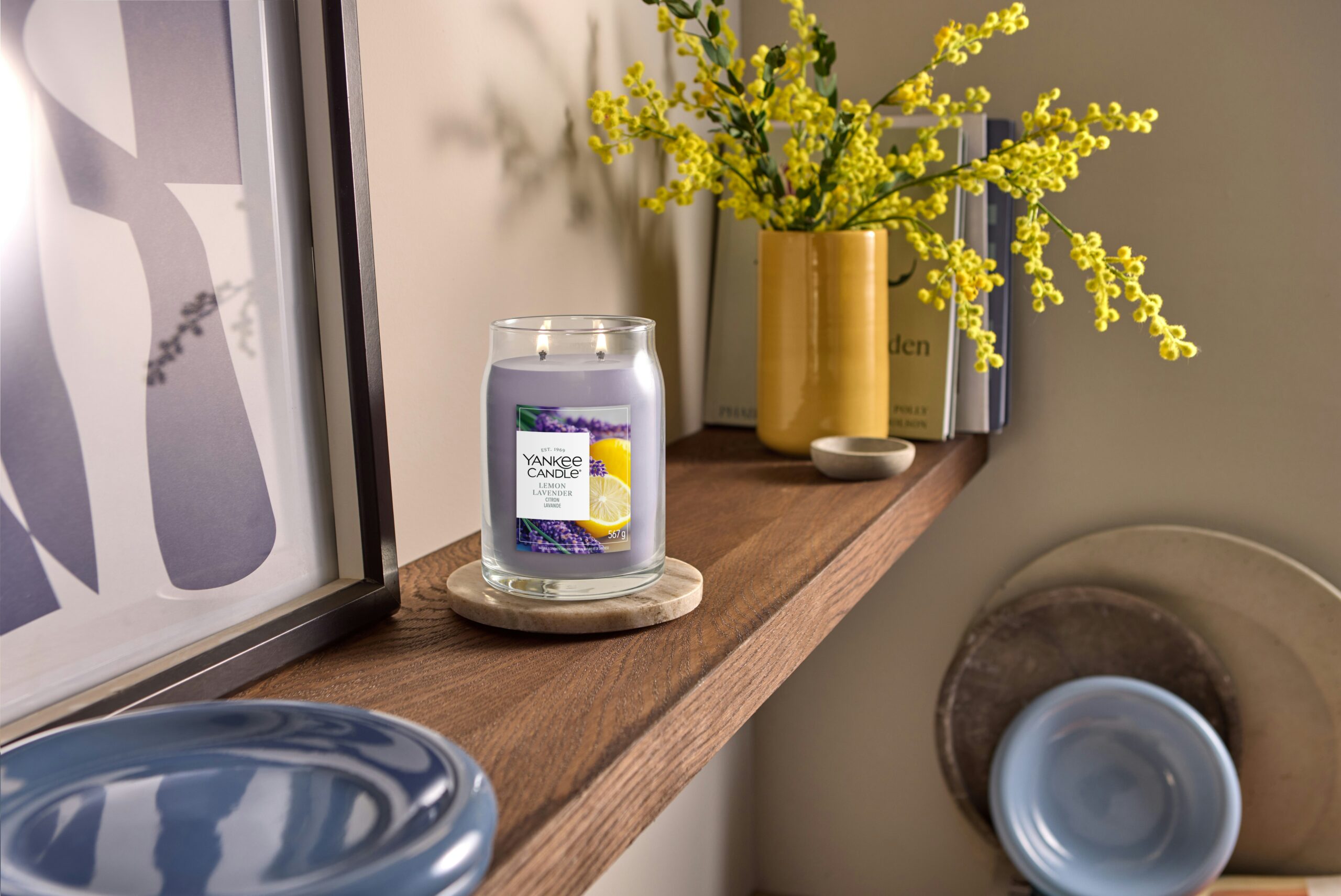 Lemon Lavender – Candela Grande – Yankee Candle 350 Punti 2 Una candela “Lemon Lavender” - Giara grande - Yankee Candle è appoggiata su una mensola di legno accanto a un vaso di fiori gialli, alcuni libri e dei piatti di ceramica blu, creando un’atmosfera accogliente e ordinata.