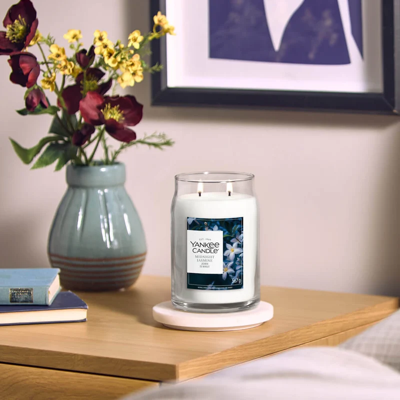 Midnight Jasmine – Giara Grande – Yankee Candle 350 Punti 5 Una candela Yankee Candle “A Midnight Jasmine - Giara Grande” di colore bianco è appoggiata su un comodino di legno accanto a un vaso di fiori, due libri e una foto incorniciata appesa alla parete, creando un'atmosfera accogliente nella camera da letto.