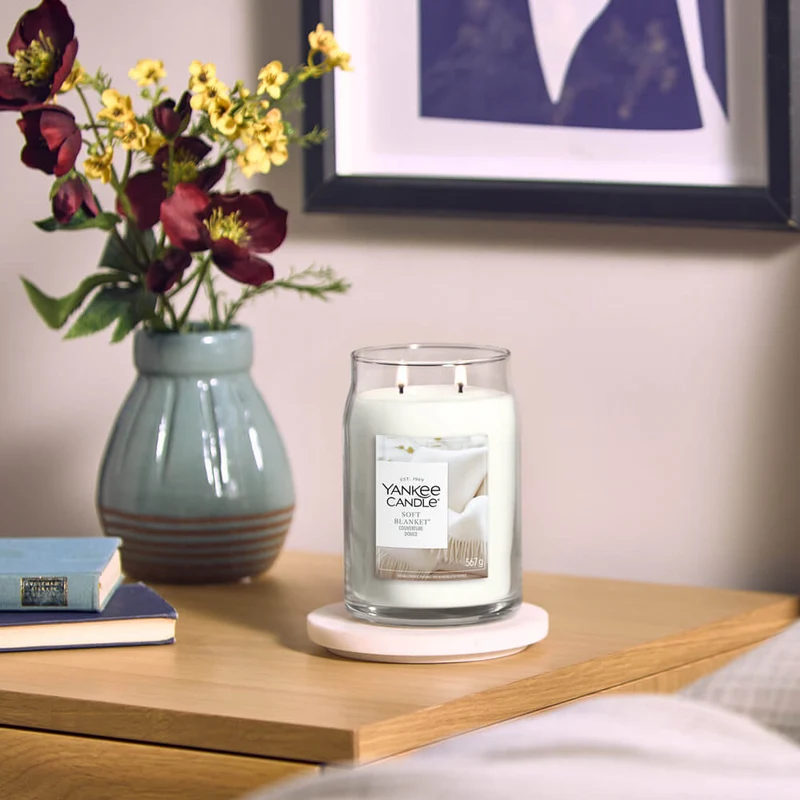 Soft Blanket – Giara Grande – Yankee Candle 350 Punti 5 Un barattolo “A Soft Blanket - Giara Grande - Yankee Candle” è posizionato su un comodino di legno accanto a due libri, una composizione floreale in un vaso verde e una foto incorniciata appesa alla parete, in un’accogliente camera da letto.