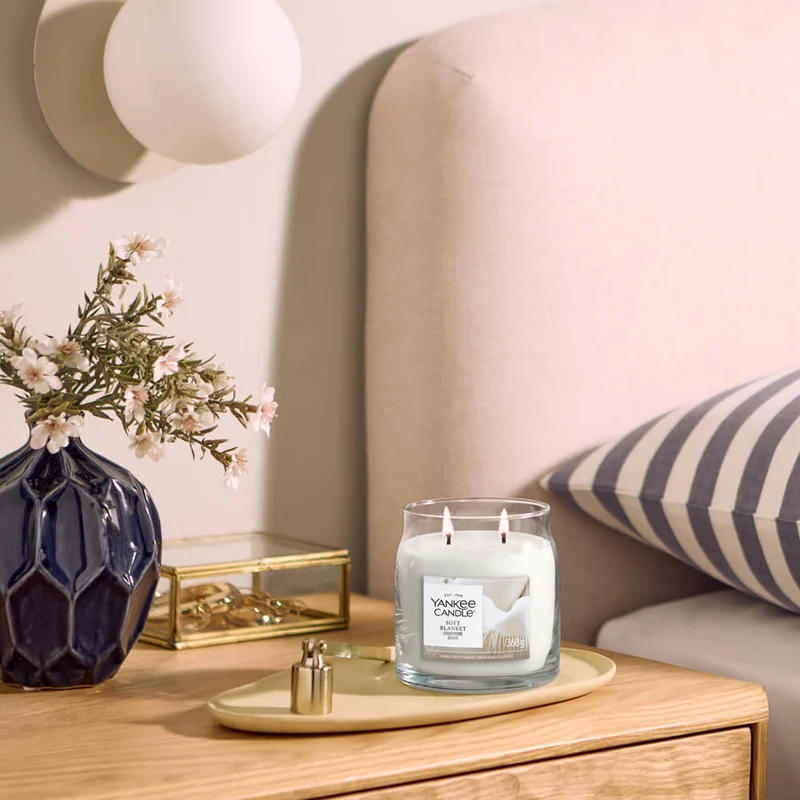 Soft Blanket – Giara Media – Yankee Candle 250 Punti 2 Un comodino con un vaso blu pieno di fiori bianchi, un portagioie di vetro e una candela accesa “Soft Blanket - Giara Media - Yankee Candle”. Sullo sfondo si intravedono una testiera beige e un cuscino a righe.