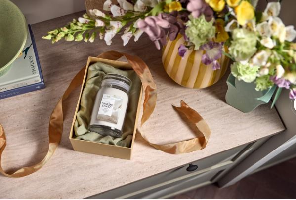 Soft Blanket – Giara Grande – Yankee Candle 350 Punti 3 Una coperta morbida - Giara Grande - Yankee Candle, racchiusa in una scatola con un nastro di raso, è appoggiata su un tavolo di legno accanto a un vaso a righe con dei fiori e a una pila di libri.