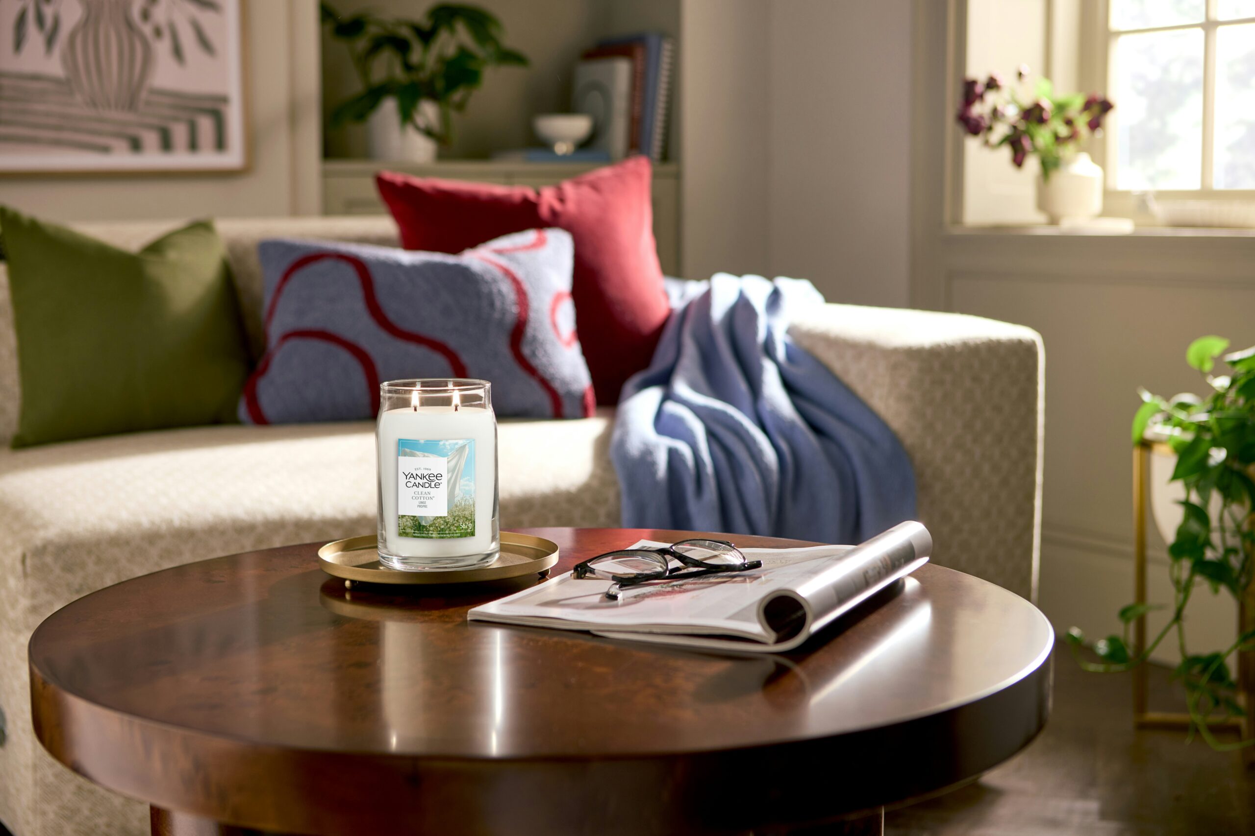Clean Cotton – Giara Grande – Yankee Candle 350 Punti 2 Un accogliente salotto con un divano bianco, cuscini colorati e una coperta blu. Sul tavolino rotondo si trova una candela Yankee Candle Clean Cotton - Giara Grande accesa, accanto a occhiali da lettura e a una rivista, mentre la luce del sole filtra dalla finestra.
