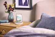 Clean Cotton – Giara Grande – Yankee Candle 350 Punti 3 Un comodino contiene una candela Clean Cotton - Giara Grande - Yankee Candle, un vaso con fiori rossi e gialli, un libro chiuso e si trova accanto a un letto con cuscini blu e grigi. Sulla parete sovrastante è appesa un'opera d'arte incorniciata.