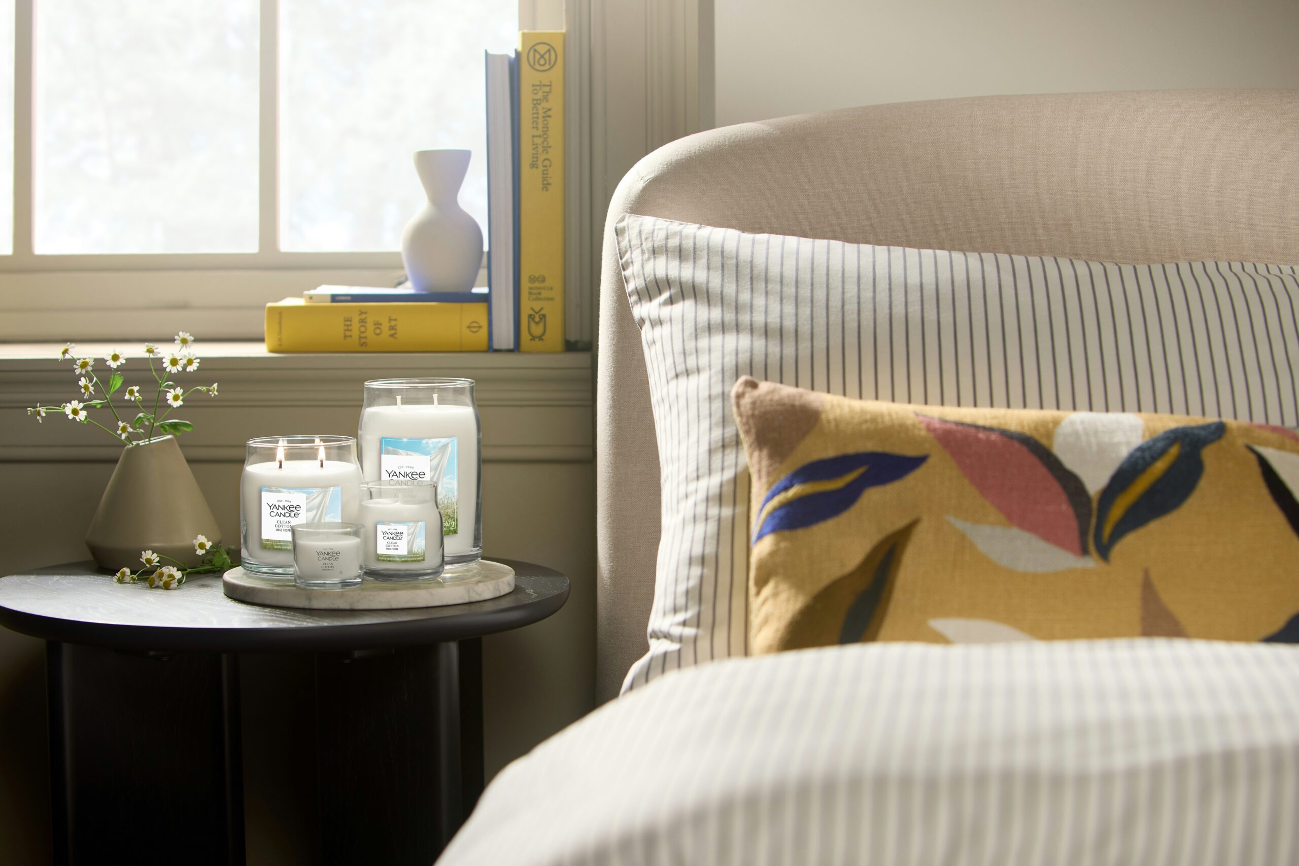 Clean Cotton – Candela Piccola – Yankee Candle 100 Punti 2 Un'accogliente camera da letto con un letto, un cuscino a righe, un cuscino giallo e un tavolino nero con tre barattoli Yankee Candle Clean Cotton Tumbler Piccolo, mentre sul davanzale della finestra si trovano dei libri alla luce del sole.