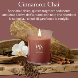 Una candela “Cinnamon Chai” di Giara Media - WoodWick è circondata da bastoncini di cannella, chiodi di garofano, sassolini e un fiore di orchidea bianca. Il testo in italiano sopra descrive le note aromatiche della candela.