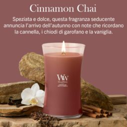 Una candela Cinnamon Chai - Candela Grande - WoodWick accesa è appoggiata su una superficie di legno, circondata da bastoncini di cannella, chiodi di garofano, un'orchidea bianca e sassi, con sopra una descrizione della fragranza in italiano.
