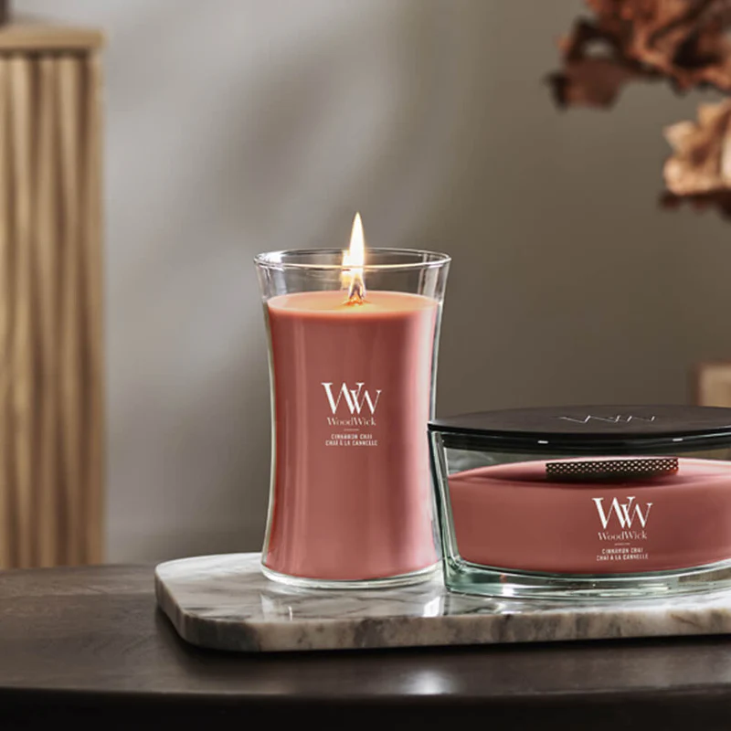 Cinnamon Chai – Candela Grande – WoodWick Giara Grande 2 Una candela WoodWick rosa accesa, contenuta in un barattolo di vetro, è appoggiata su un vassoio di marmo accanto a una candela WoodWick "Cinnamon Chai - Giara Grande" spenta e con il coperchio. Sullo sfondo si intravedono le calde luci dell'interno e delle foglie autunnali sfocate.