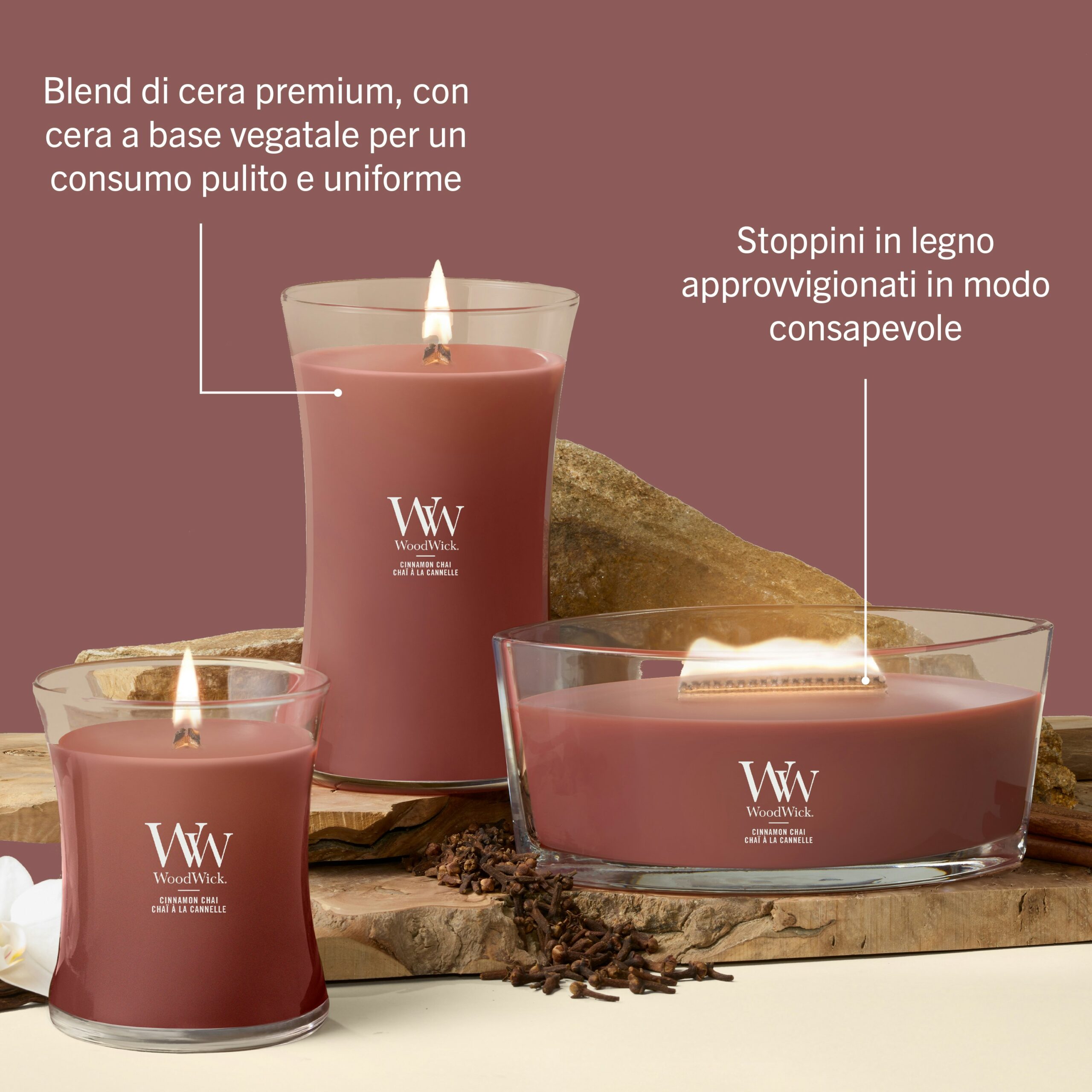 Cinnamon Chai – Candela Grande – WoodWick Giara Grande 4 Tre candele Cinnamon Chai - Candela Grande - WoodWick, caratterizzate da una ricca tonalità rosso-marrone, sono accese ed esposte su una superficie beige impreziosita da pietre e legno. Il testo in italiano mette in risalto le caratteristiche principali, quali la miscela di cera di alta qualità, l'invitante fragranza Cinnamon Chai e gli stoppini in legno provenienti da fonti responsabili.