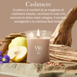 Una candela “Lit Cashmere” di Giara Media - WoodWick è disposta tra un frutto di mela cotogna, legno di sandalo, un fiore viola e pezzi di legno, con un testo in italiano che ne descrive le note olfattive calde e cremose.