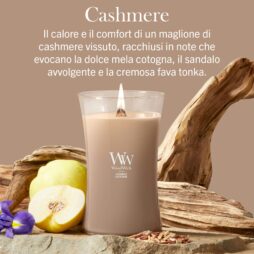 La candela Cashmere - Giara Grande - WoodWick è accesa, circondata da frutti di mela cotogna, legno di sandalo, fave tonka e fiori viola su uno sfondo beige, con una descrizione in italiano sopra.