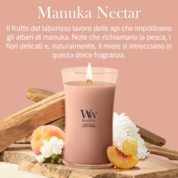 Una candela “A Manuka Nectar - Giara Grande - WoodWick” è esposta accanto a elementi decorativi in legno, una pesca e fiori bianchi, con una didascalia in italiano che ne sottolinea la dolce fragranza ispirata al miele, alla pesca e a delicate note floreali.