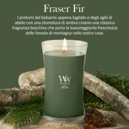 Una candela Frasier Fir - Giara Grande - WoodWick, con la sua tonalità verde e il caratteristico profumo di pino, è accesa e posizionata su un tavolo decorato con rami di pino, pigne e resina; accanto, un testo in italiano ne descrive l’aroma fresco e boschivo.
