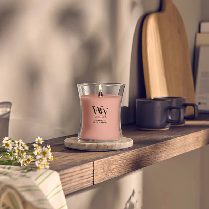 Manuka Nectar – Giara Media – Woodwick Giara media 2 Una candela “Manuka Nectar” di Giara Media con stoppino in legno è accesa e poggia su una mensola di legno sopra un sottobicchiere di marmo, circondata da una tazza nera, un tagliere, un libro e un piccolo mazzo di fiori bianchi. Una luce calda e naturale riempie lo spazio creando un’atmosfera accogliente.