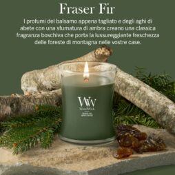 Una candela WoodWick “Frasier Fir - Giara Media”, con la fiammella verde accesa, poggia su tronchetti di betulla, affiancata da pezzi di resina. Il testo in italiano sopra evidenzia la sua fragranza boschiva, ambrata e di abete. L’ambientazione naturale e gli oggetti evocano l’atmosfera di un bosco.