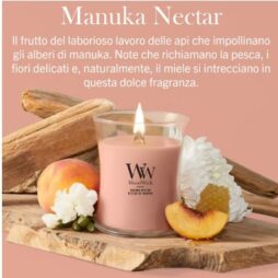 Una candela “A Manuka Nectar” - Giara Media - Woodwick, di colore rosa, poggia su una base di legno, circondata da fiori bianchi, fette di pesca e favi di miele, su uno sfondo color pesca con un testo in italiano che descrive la fragranza.
