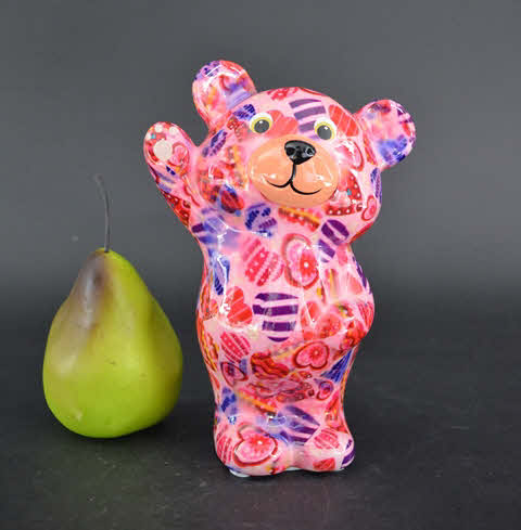 Bear Tilou- Colore Rosa – Pomme Pidou Bear L'orso Tilou - Colore Rosa - Pomme Pidou, un colorato orsetto di ceramica con motivi rosa, rossi e viola che agita una zampa, è in piedi accanto a una pera verde su uno sfondo nero.