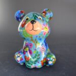 Il Pomme Pidou Teddy Bear Tala presenta un design in ceramica verde con motivi floreali colorati, tra cui fiori arancioni, gialli e rossi. Mostra occhi grandi e un sorriso discreto mentre è seduto dritto su una superficie scura.