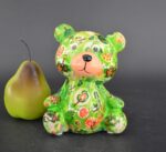 Un orsetto decorativo Toto di Pomme Pidou in Colore Verde, caratterizzato da motivi floreali rossi, arancioni e bianchi, è posizionato in posizione verticale accanto a una pera verde con gambo scuro su uno sfondo scuro e semplice.