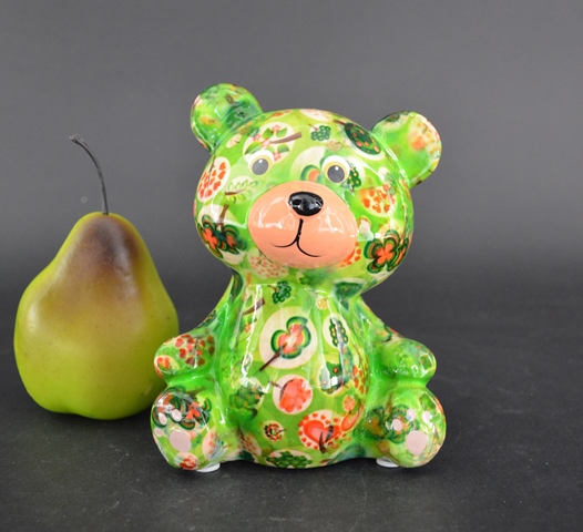 Teddy Bear Toto – Colore Verde – Pomme Pidou Bear Un orsetto decorativo Toto di Pomme Pidou in Colore Verde, caratterizzato da motivi floreali rossi, arancioni e bianchi, è posizionato in posizione verticale accanto a una pera verde con gambo scuro su uno sfondo scuro e semplice.