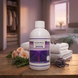 Un flacone di Profuma Bucato - Lavanda Aromatica - Horomia da 500 ml è appoggiato su un tavolo di legno con asciugamani ripiegati, lavanda e fiori, mentre un letto ben fatto e un'illuminazione delicata esaltano l'atmosfera accogliente della camera da letto sullo sfondo.