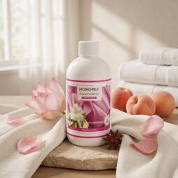 Una bottiglia da 500 ml di Horomia Muschio e Loto Profuma Bucato è appoggiata sul legno, circondata da fiori di loto rosa, anice stellato, pesche fresche e asciugamani bianchi ripiegati in un ambiente tranquillo, simile a una spa.