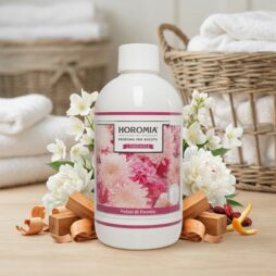 Una bottiglia da 500 ml di Profuma Bucato Horomia "Petali di Peonia" si trova tra fiori di peonia, bastoni di legno e cesti di biancheria su una superficie di legno, con asciugamani bianchi ordinatamente piegati sullo sfondo.
