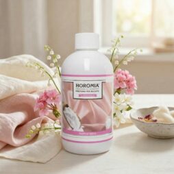 Una bottiglia da 500 ml di Soffice Talco - Profuma Bucato Horomia è appoggiata su un tavolo ornato di fiori bianchi e rosa, accanto a un panno beige e rosa e a una ciotola di petali, immersa in una luce naturale soffusa per un'atmosfera invitante.