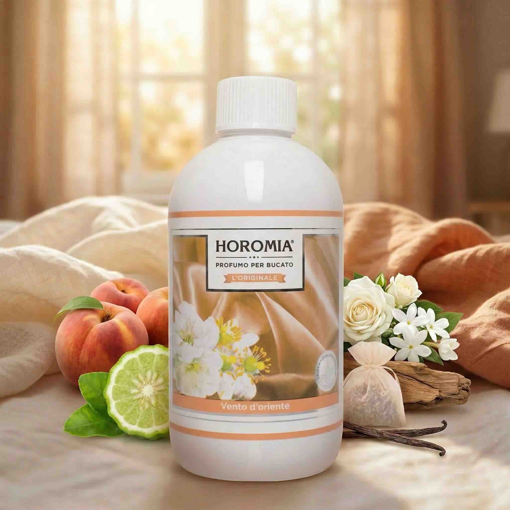 Vento D’Oriente – Profuma Bucato – Horomia 500 ml Horomia 500 ml Una bottiglia di Vento D'Oriente - Profuma Bucato - Horomia 500 ml è appoggiata su fogli beige, circondata da fiori freschi, pesca, bergamotto e baccello di vaniglia, creando un ambiente morbido e accogliente.