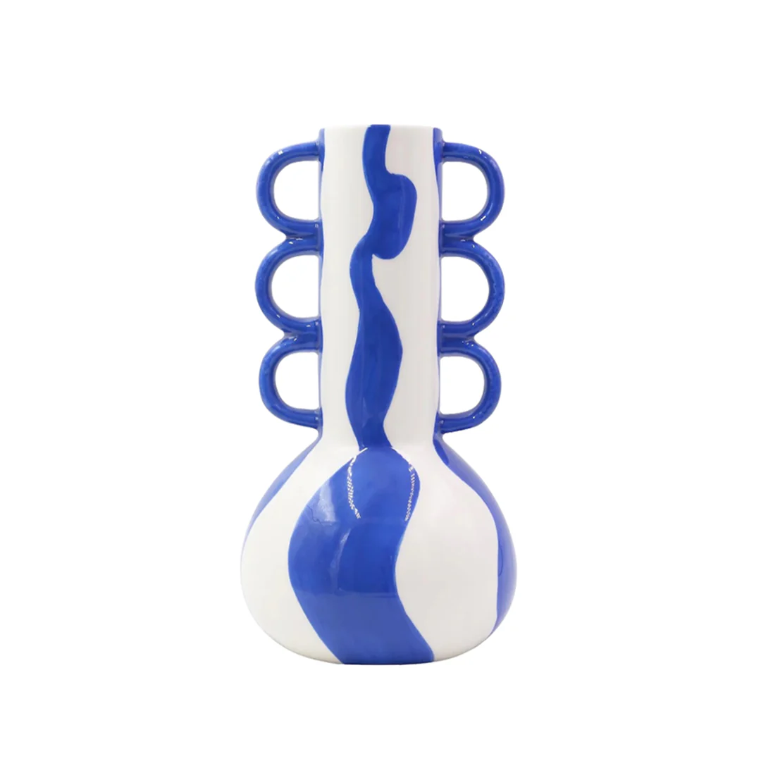 Tomas Vaso in ceramica – Que Rico Querico Il Tomas Vaso in ceramica - Que Rico è un vaso in ceramica bianca caratterizzato da una base bulbosa, strisce ondulate blu verticali e tre manici ad anello blu su ciascun lato.