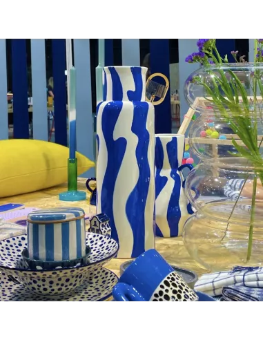 Vaso Esteban in ceramica – Que Rico Querico 2 Un vivace allestimento da tavolo che presenta il Vaso Esteban in ceramica di Que Rico, vasi e piatti Que Rico a righe blu e bianche e a pois, un cuscino giallo, fiori e oggetti decorativi colorati sullo sfondo.