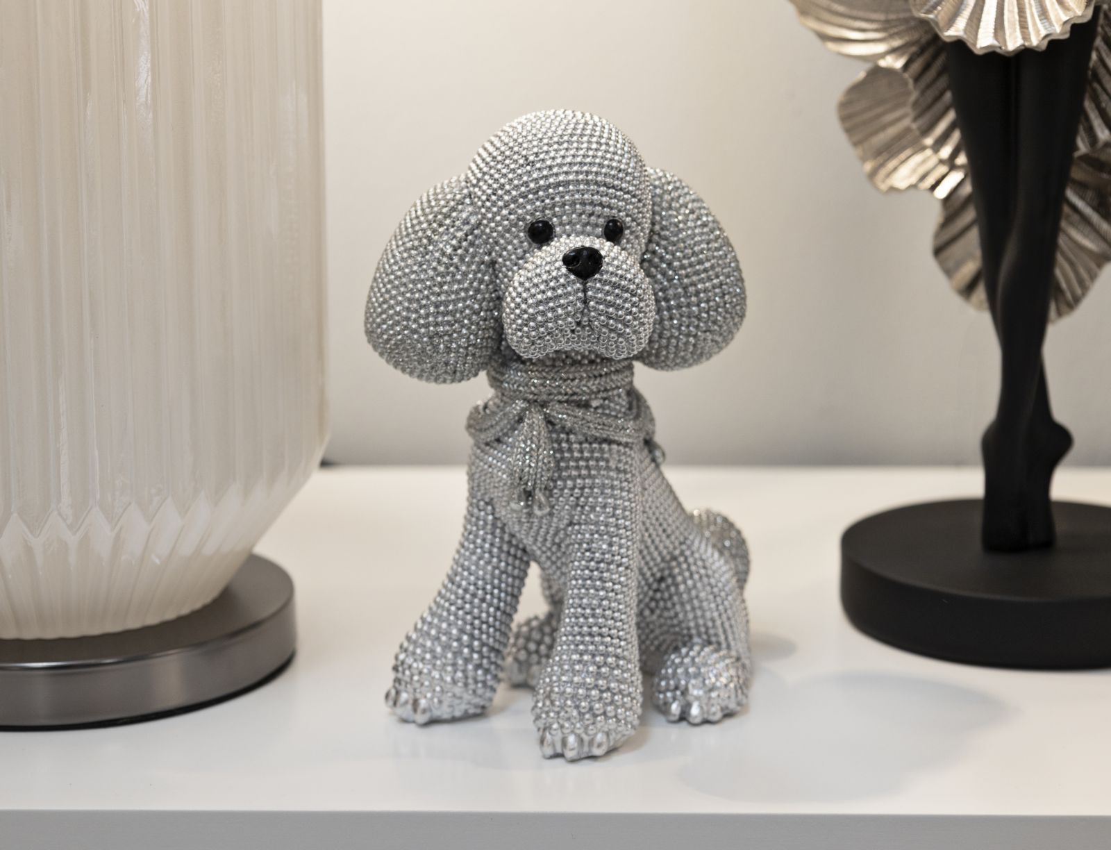 Barboncino Sparky – Mauro Ferretti – 15 x 15 x 9.5 cm Figure 3 La statuina Barboncino Sparky di Mauro Ferretti, decorata con strass scintillanti, è esposta su una mensola bianca tra sculture bianche e nere.