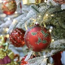 Primo piano di un ramo dell'albero di Natale decorato con aghi smerigliati, con un ornamento rosso ornato da agrifoglio verde e volute dorate, circondato da luci bianche e altri ornamenti.