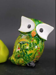 Il gufo Otti con rami - Pomme pidou in ceramica, caratterizzato da un motivo floreale e di foglie verde brillante, grandi occhi bianchi, becco arancione e zampe nere, &egrave; affiancato da un oggetto verde chiaro su sfondo scuro.