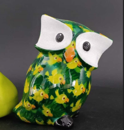 Il Gufo Otti con Pappagalli della collezione Pomme pidou &egrave; un gufo in ceramica con grandi occhi bianchi, becco arancione e decorazioni colorate a forma di pappagallo. Si trova accanto a una mela verde su un deciso sfondo nero.