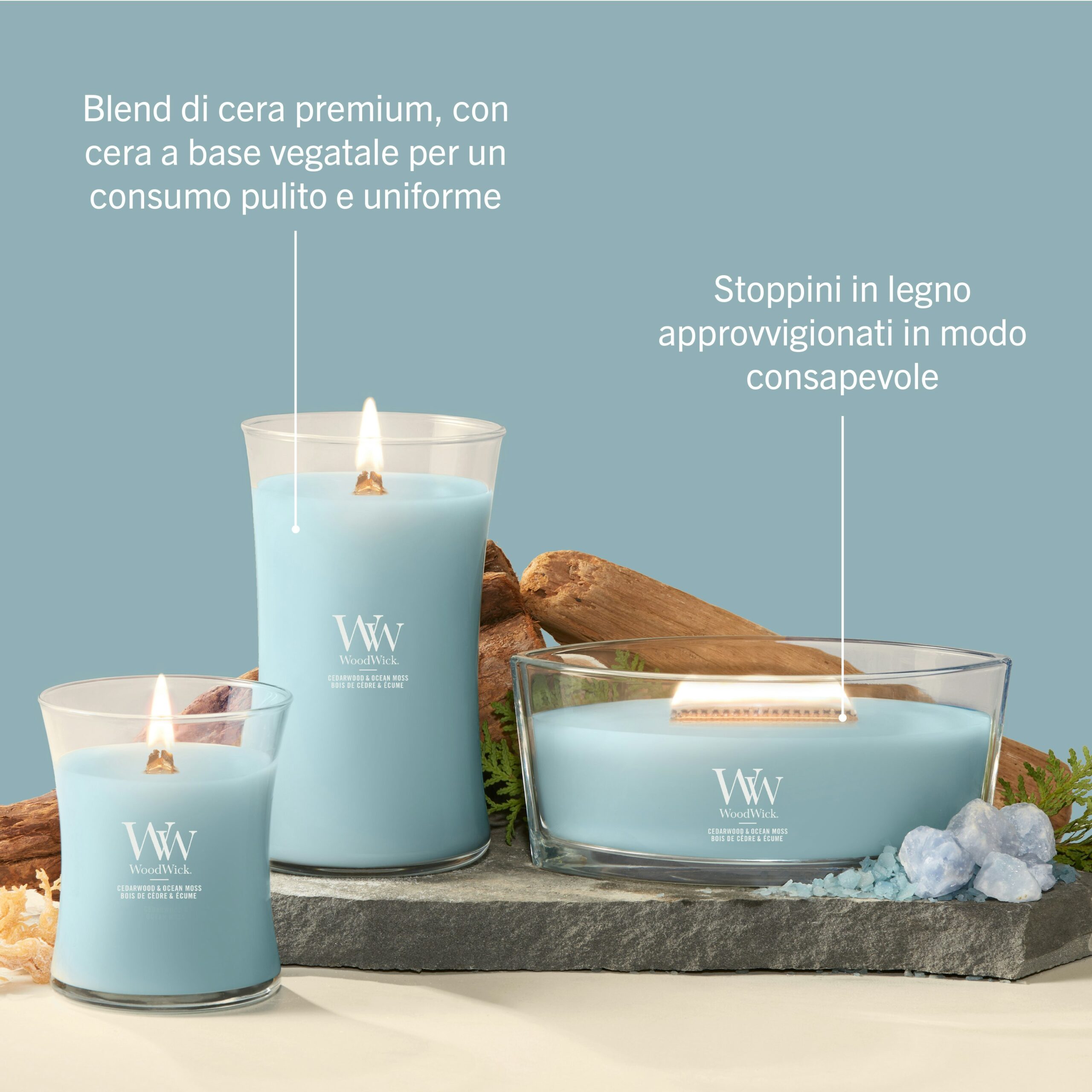 Cedarwood & Ocean Moss – Candela Grande – Woodwick Giara Grande 5 Tre candele “Cedarwood & Ocean Moss” - Candela Grande - Woodwick, contenute in vasetti di vetro blu, sono disposte su una superficie beige, circondate da pietre decorative e cristalli. Il testo in italiano sottolinea la loro miscela di cera vegetale di alta qualità, la delicata fragranza di muschio marino e legno di cedro, nonché gli stoppini in legno provenienti da fonti responsabili che garantiscono una combustione di alta qualità.