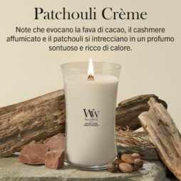 Una candela accesa della linea Patchouli Creme - Giara Grande - Woodwick è appoggiata su una superficie di pietra, circondata da legno, fave di cacao e foglie secche, mentre il testo in italiano sopra descrive la sua fragranza.