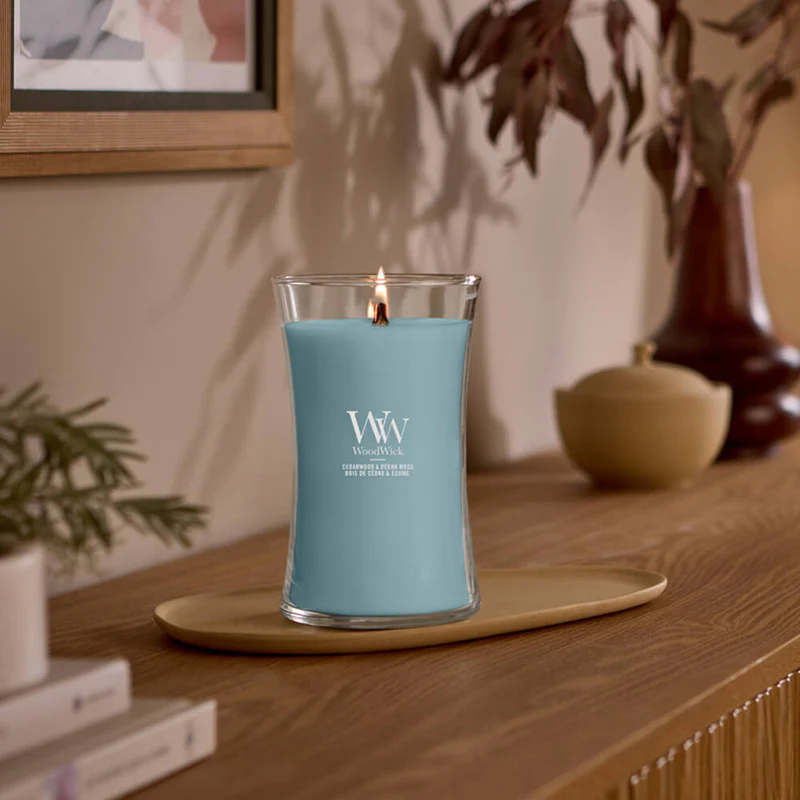 Cedarwood & Ocean Moss – Candela Grande – Woodwick Giara Grande 2 Una Candela Grande di WoodWick al legno di cedro e muschio oceanico brucia su un vassoio ovale di legno su una superficie di legno, con decorazioni e piante sfocate sullo sfondo, creando un'atmosfera accogliente e invitante.