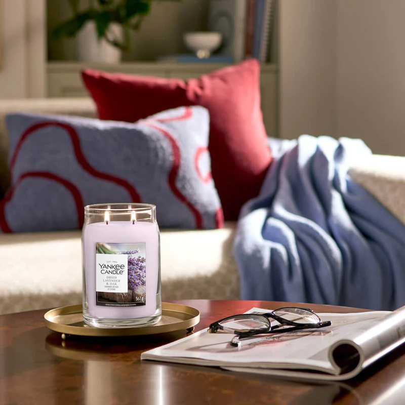 Dried Lavender And Oak – Candela Grande – Yankee Candle 350 Punti 2 Una candela Yankee Dried Lavender & Oak - Candela Grande accesa poggia su un vassoio con una rivista e degli occhiali sul tavolino; sullo sfondo, un divano con cuscini rossi e grigi e una coperta blu.