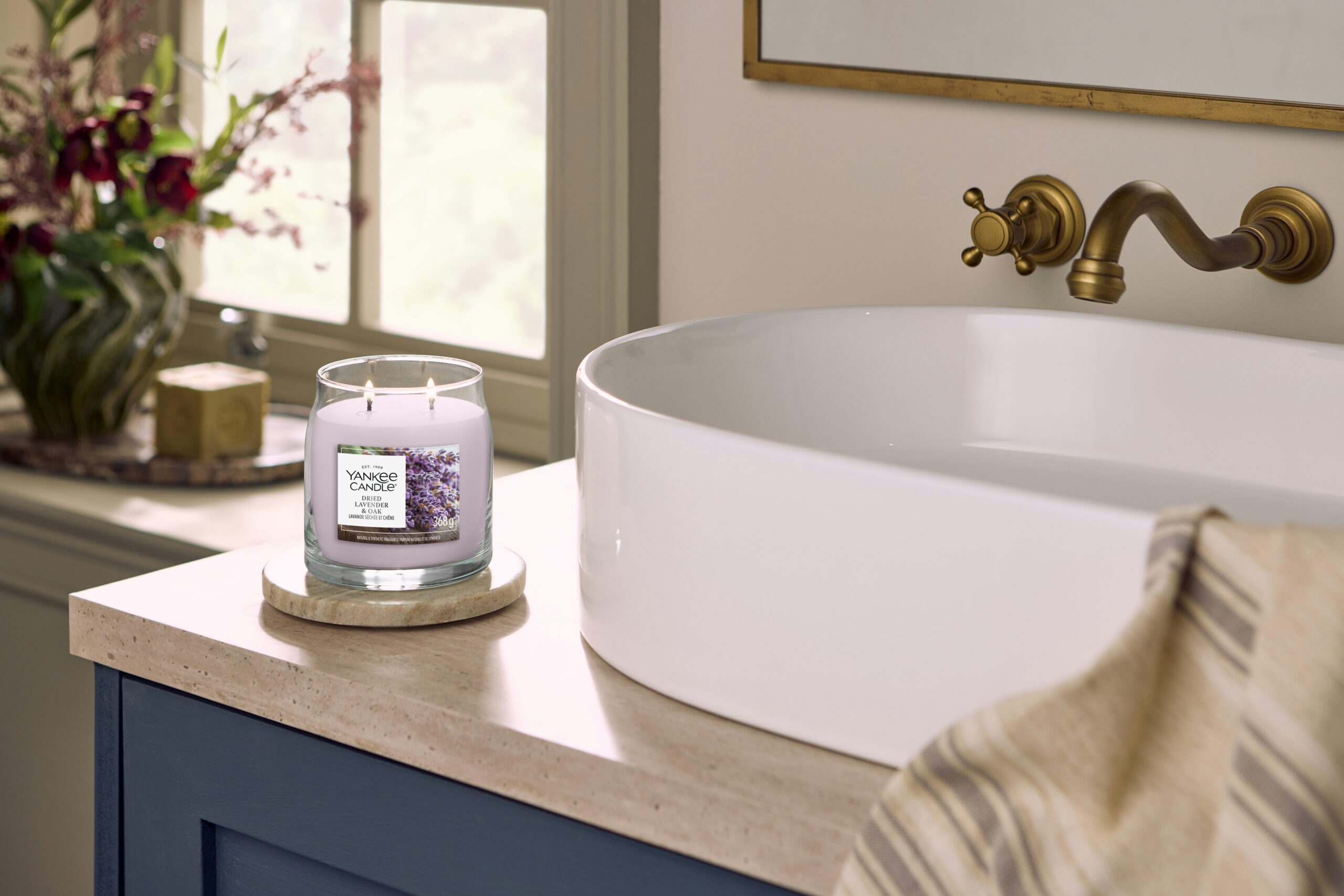 Dried Lavender And Oak – Candela Media – Yankee Candle 250 Punti 2 Una candela Yankee Dried Lavender & Oak - Candela Grande brucia su un sottobicchiere vicino a un moderno lavandino bianco, riempiendo il luminoso bagno - caratterizzato da un rubinetto dorato e da un asciugamano a righe - con note rilassanti di lavanda secca.