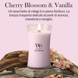 Una candela rosa “Cherry Blossom And Vanilla - Candela Grande - WoodWick” in un barattolo di vetro è appoggiata su una superficie di legno e pietra, circondata da fiori di ciliegio. L’etichetta riporta il nome del prodotto, mentre il testo sopra descrive la fragranza come una miscela di note di arancia, mandorla e vaniglia cremosa che racchiude la delicata fragranza dei fiori di ciliegio.