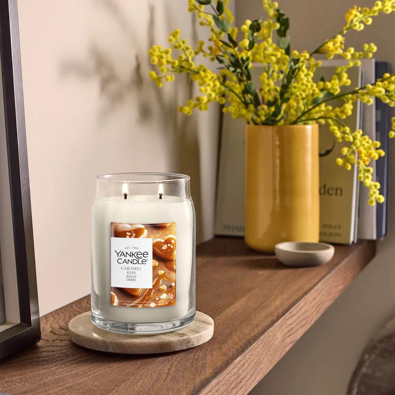 Caramel Kiss – Candela Grande – Yankee Candle 350 Punti 2 Una Yankee Candle Caramel Kiss - Candela Grande con due stoppini si trova su un sottobicchiere di marmo su una mensola di legno, con libri e un vaso di fiori gialli disposti dietro di essa.