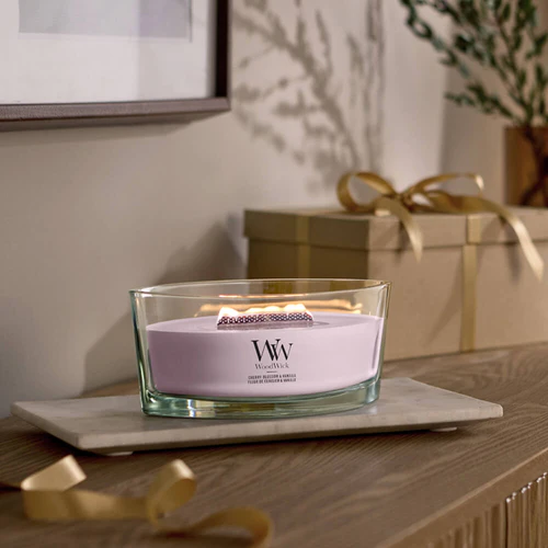 Cherry Blossom And Vanilla – Ellipse – Woodwick Ellipse 2 Una candela WoodWick Ellipse Cherry Blossom And Vanilla brucia su una superficie di legno, riempiendo l'aria di un dolce profumo. Un nastro dorato e una confezione regalo sullo sfondo creano un'atmosfera accogliente.