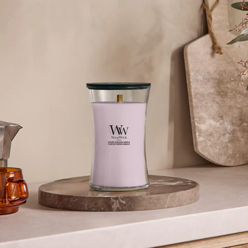 Cherry Blossom And Vanilla – Candela Grande – Woodwick Giara Grande 2 La candela Cherry Blossom And Vanilla - Candela Grande - Woodwick, una candela bianca con coperchio nero, si trova su un vassoio rotondo in pietra su un piano di lavoro beige, riempiendo la stanza con una fragranza rilassante. Sullo sfondo sono visibili un tagliere e una decorazione.
