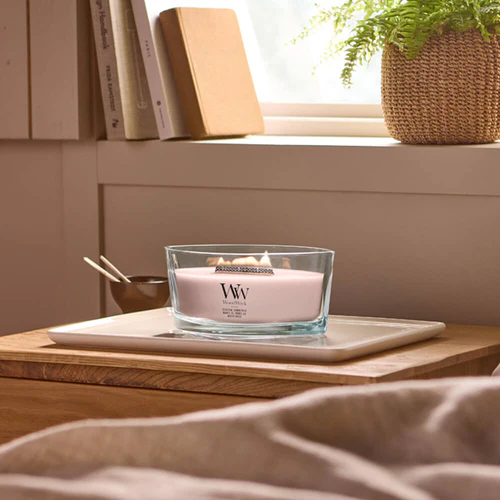 Coastal Sunberry – Ellipse – Woodwick Ellipse 2 Sul comodino in legno bianco è accesa una candela Coastal Sunberry - Ellipse di Woodwick, posta su un computer portatile chiuso. Sul davanzale della finestra, sullo sfondo, sono visibili una piccola pianta e dei libri.