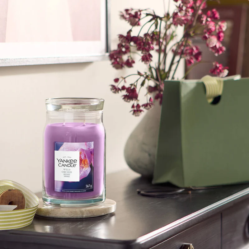 Wild Orchid – Candela Grande – Yankee Candle 250 Punti 2 Una candela Wild Orchid - Candela Grande - Yankee Candle si trova su un sottobicchiere rotondo su un comò scuro, con un vaso di fiori viola, una borsa della spesa verde e parte di una foto incorniciata visibili dietro di essa.